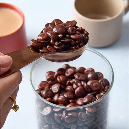 Spoonful of chocolate chips over a glass container with more chocolate chips, on a light blue background.