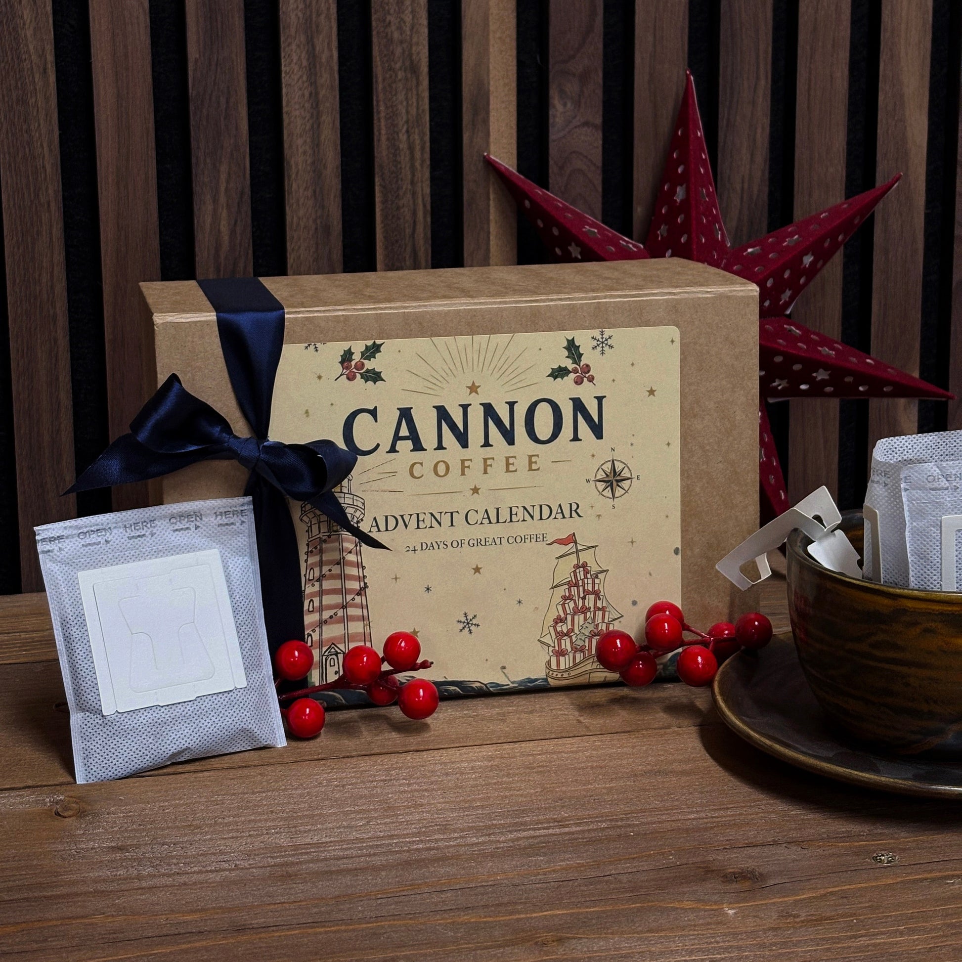 Cannon Coffee Advent Calendar gift box in kraft brown with a navy ribbon, displayed on a wooden table with coffee bags, red berries, a cup, and a red star ornament against a dark wood slat background.
