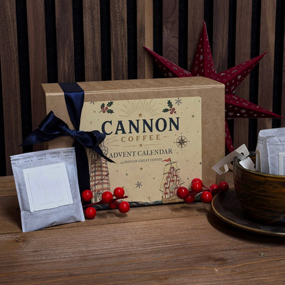 Cannon Coffee Advent Calendar gift box in kraft brown with a navy ribbon, displayed on a wooden table with coffee bags, red berries, a cup, and a red star ornament against a dark wood slat background.