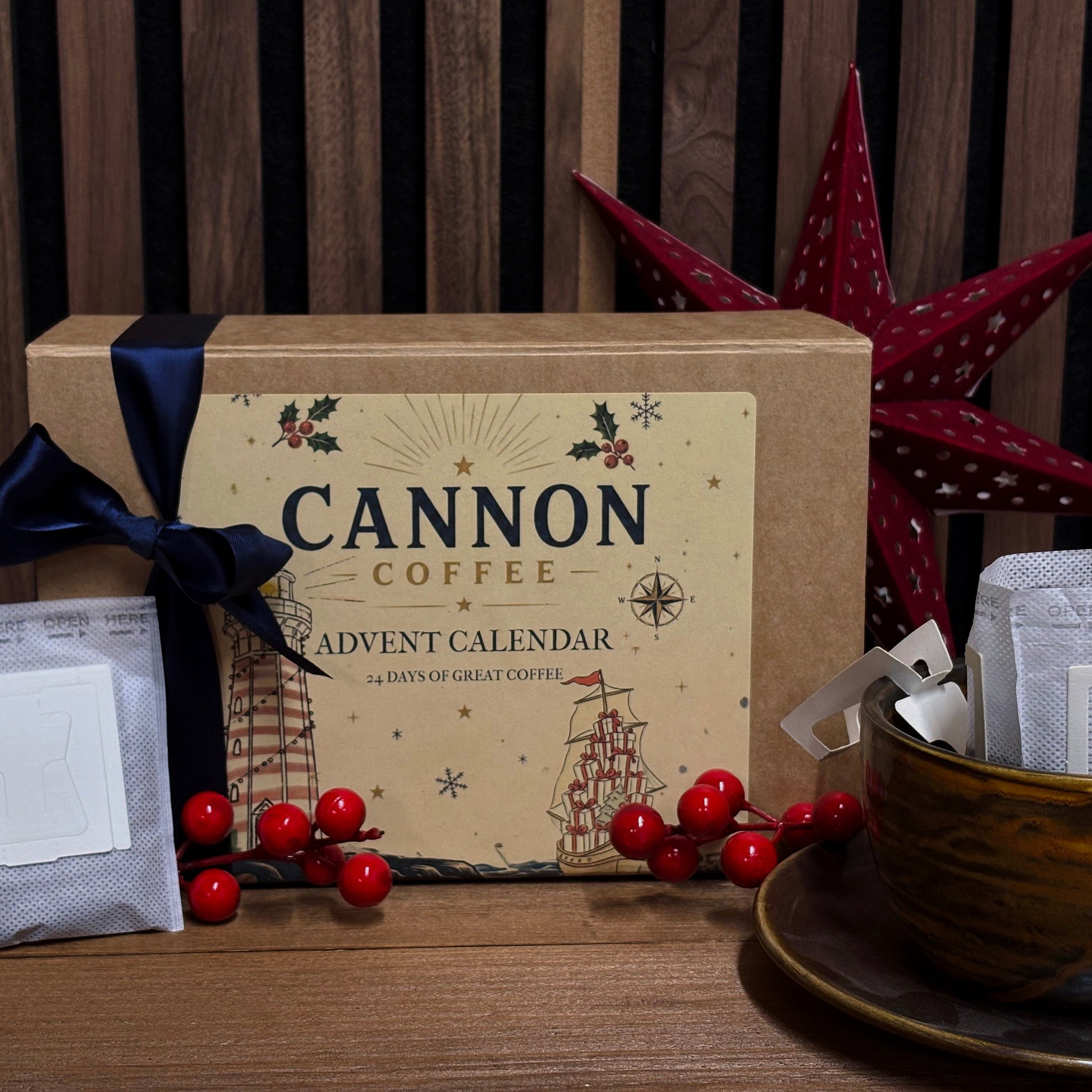 Cannon Coffee Advent Calendar gift box with a kraft paper label, navy ribbon, coffee bags, and a ceramic mug on a wooden table against a wood slat wall.