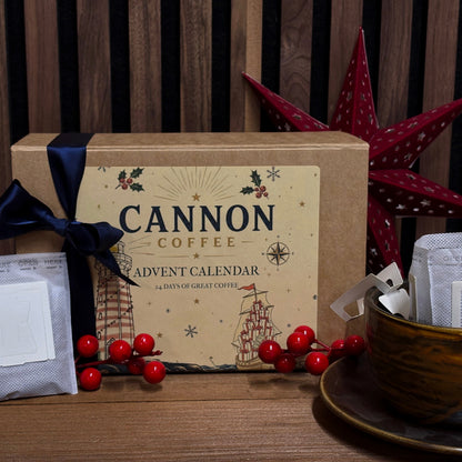 Cannon Coffee Advent Calendar gift box with a kraft paper label, navy ribbon, coffee bags, and a ceramic mug on a wooden table against a wood slat wall.