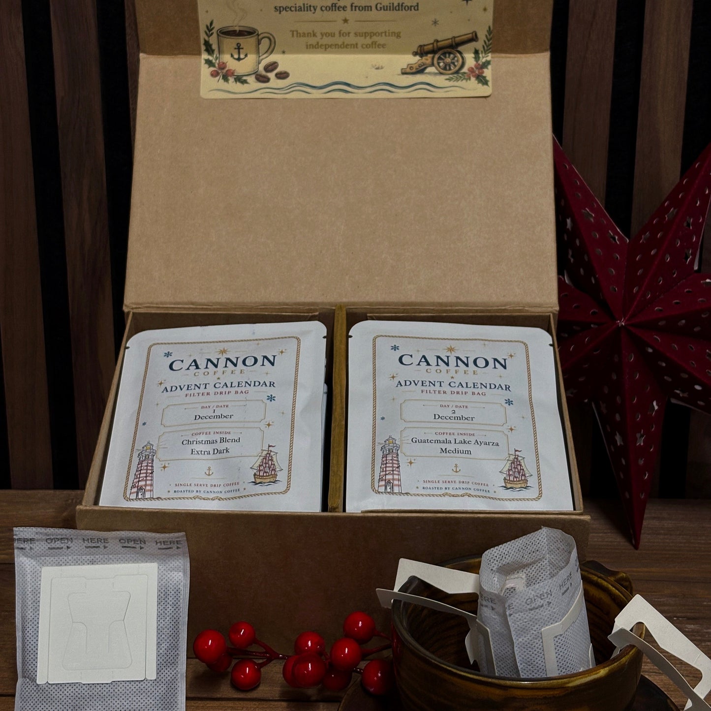 An open kraft gift box containing two boxed 'CANNON Advent Calendar' coffee packages, with additional coffee sachets and a small bowl of red berries on a table in front. The box lid has a decorative insert card, and the background shows dark wood paneling with a red star ornament.