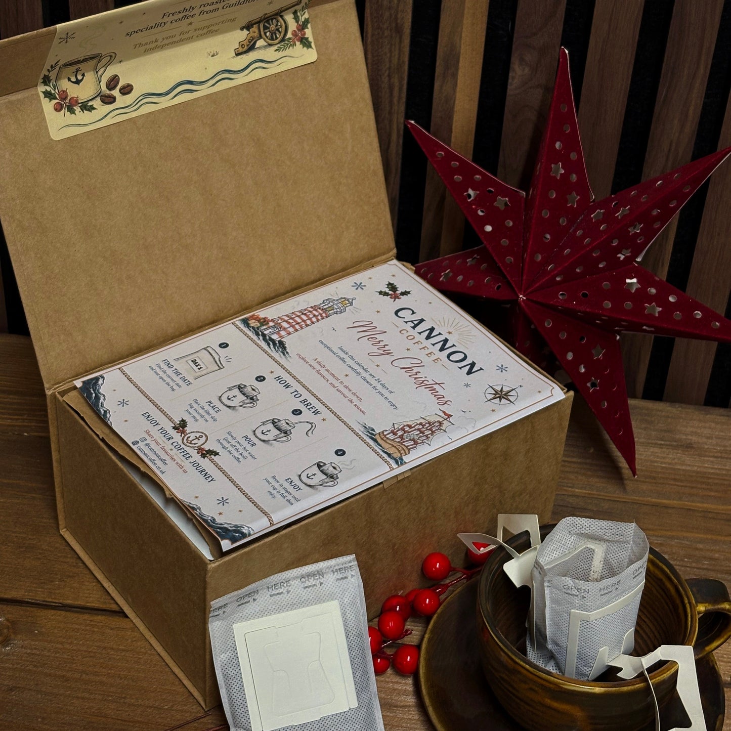 A kraft paper gift box opened to reveal printed cards and tea bags, styled on a wooden surface with a red star decoration, red berries, and a teacup.