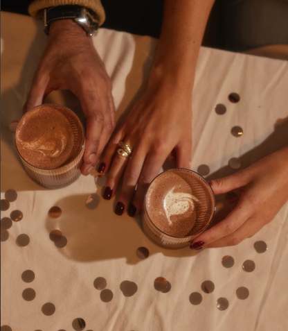 Two hands holding small cups of iced coffee or latte on a beige tablecloth scattered with metallic confetti. The scene is warmly lit with a cozy, casual aesthetic.