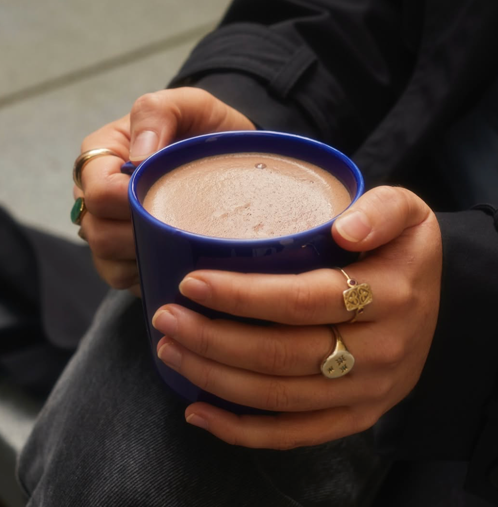 A close-up of a person holding a dark blue ceramic mug filled with a frothy hot chocolate or coffee, wearing gold rings on their fingers.