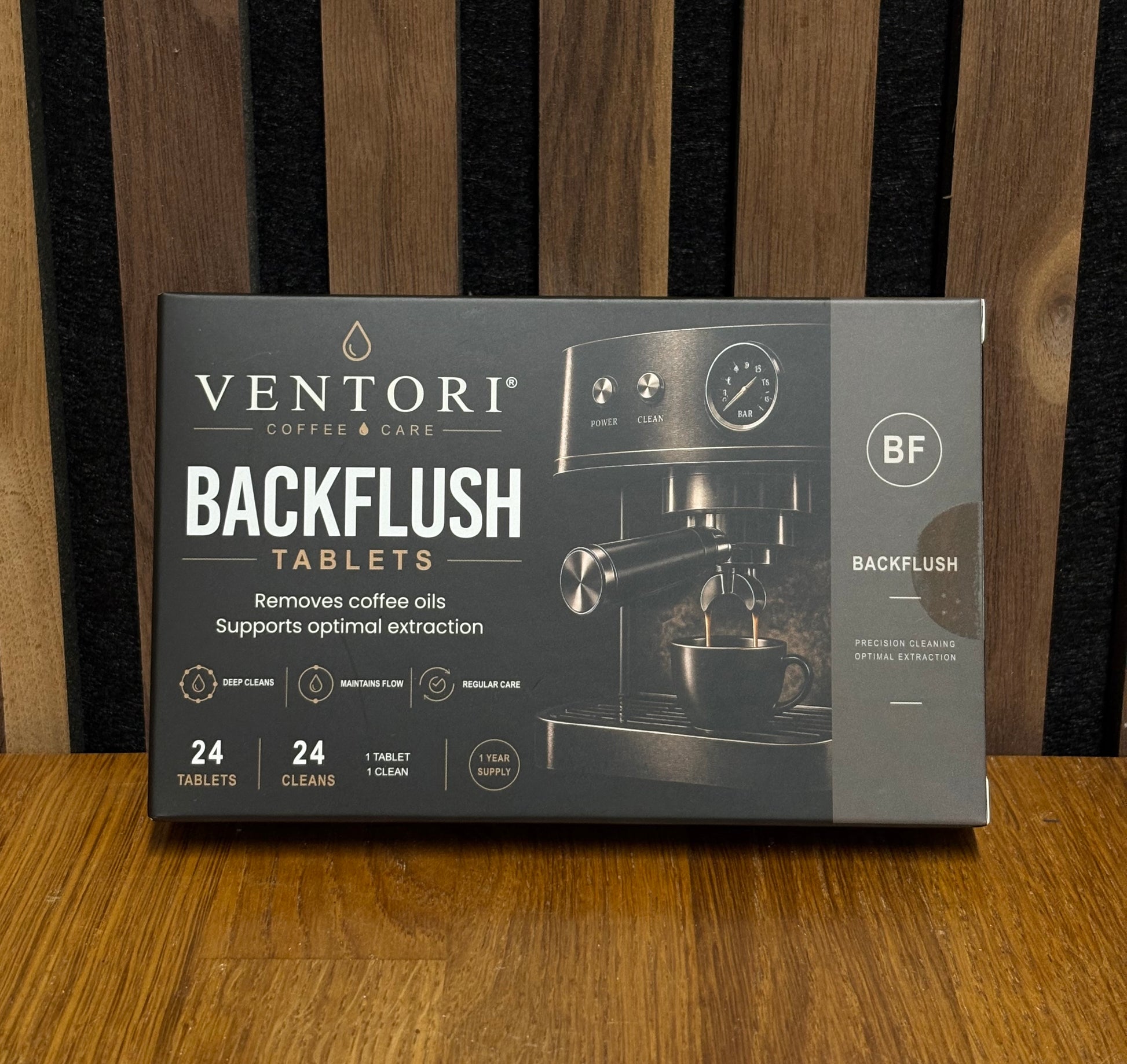 A box of Ventori Coffee & Care Backflush Tablets, featuring a dark gray and black design with an illustration of a coffee machine, placed on a wooden surface against a dark wood slatted background.